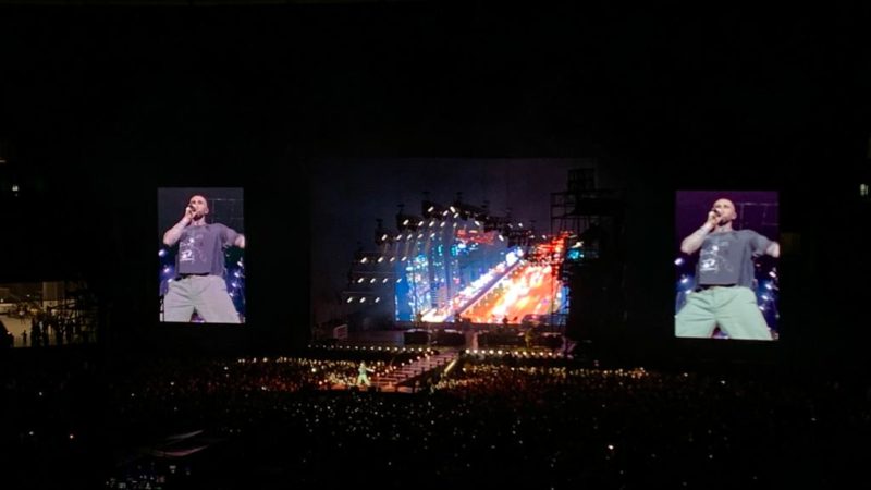 Adam Levine, vocalista da Maroon 5, em destaque em dois telões ao lado de um palco com luzes nas cores azul e vermelho.
